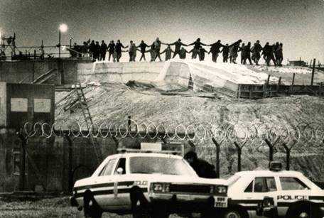 Black and white photograph shows persons performing a circle dance above silos. Two police cars are seen in the foreground.