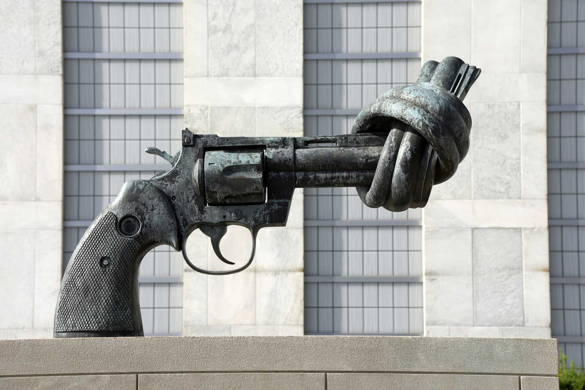 Large metal sculpture of a revolver with a knot in the barrel is seen against a stone building facade.