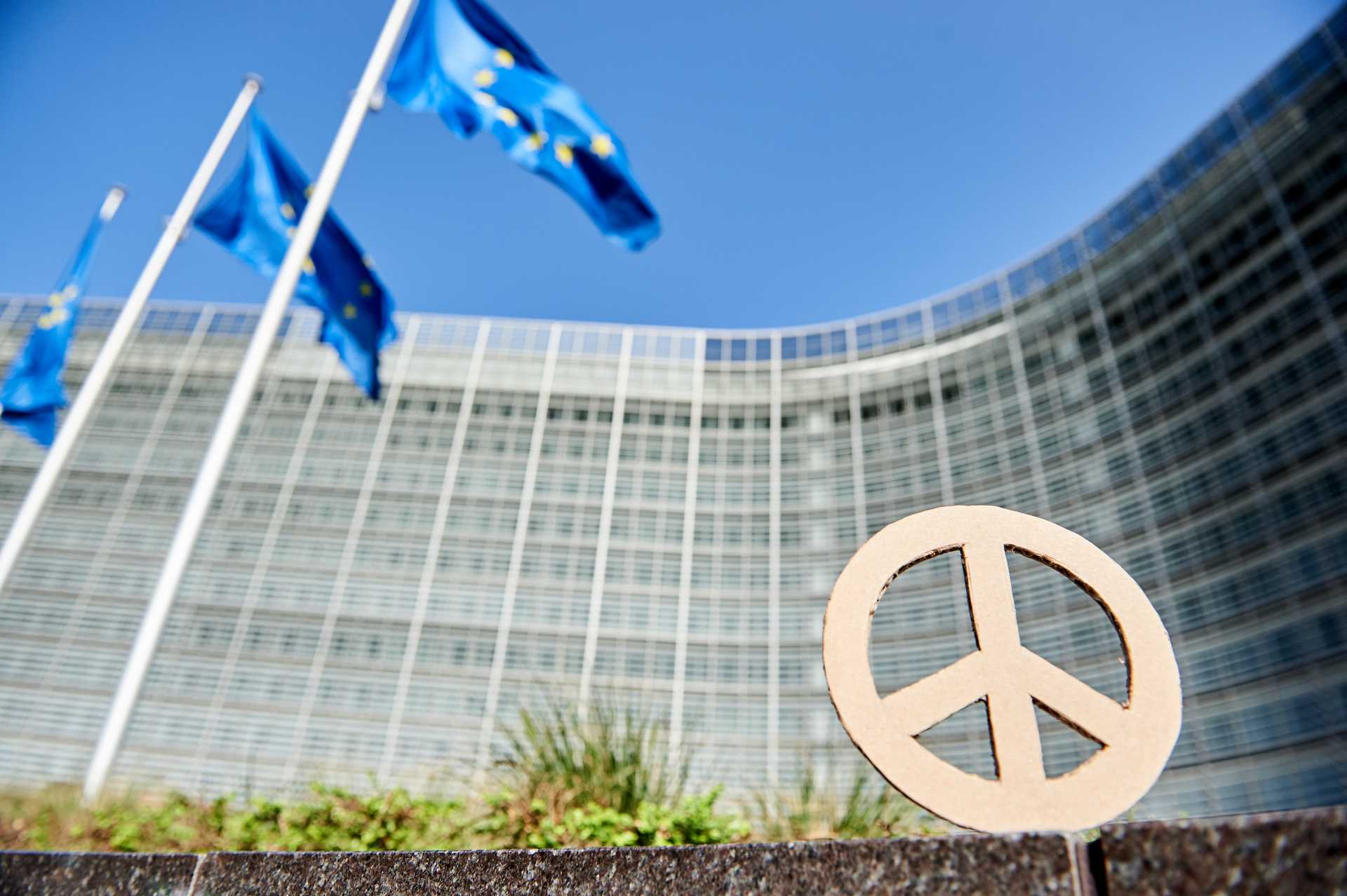 A cardboard peace symbol is seen against a looming office block, European flags, and a blue sky.